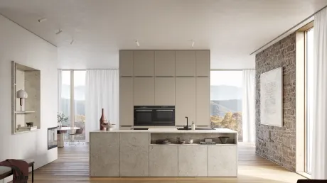 Cuisine spacieuse et moderne avec grand îlot en pierre claire et placards crème. En arrière-plan, baies vitrées sur paysage montagneux automnal. À droite, mur en pierre rustique avec œuvre d'art ; à gauche, cheminée et coin repas.