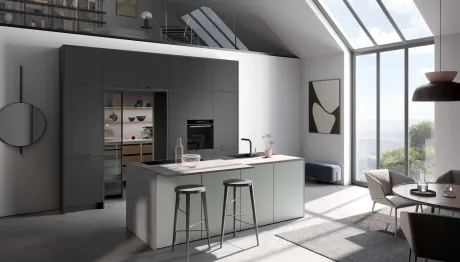 Cuisine lumineuse et moderne avec un îlot central vert clair et des placards anthracite. Îlot avec plan de travail gris, évier et deux tabourets de bar foncés. Grandes fenêtres et lucarnes sur des collines. À droite, une table à manger.