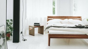Minimalistisches Schlafzimmer in Weiß mit Holzbett, Nachttisch mit Büchern und Pflanze. Licht fällt durch ein Fenster mit weißen Vorhängen. Ein gerahmtes Porträt ist im Hintergrund zu sehen.