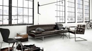 Ein loftartiger Raum mit großer Fensterfront, braunem Ledersofa und Sessel. Eine schmale Stehlampe hängt an der Wand. Ein schwarzer Stuhl und ein Teppich vervollständigen die Einrichtung.