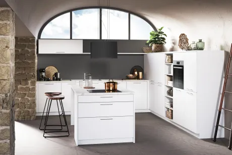 Cuisine moderne avec placards blancs et murs sombres, dominée par une grande fenêtre en arc sur le ciel. Îlot blanc avec plaque et casserole en cuivre, deux tabourets en bois. Mur en pierre à gauche, étagères à droite.