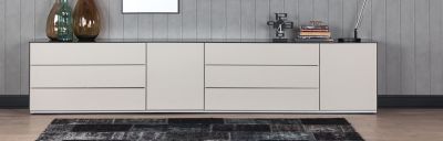 Lange, helle Kommode mit Schubladen und Schränken. Oben drauf Vasen, Uhr, Bücher und Lampe. Davor ein dunkler Teppich. Graue, vertikale Wandpaneele im Hintergrund.