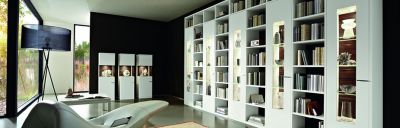 Modernes Interieur: Heller Sessel, Stehleuchte, Bücherregal mit Deko-Objekten, Vitrinen und ein Ausblick durch ein großes Fenster auf Natur.