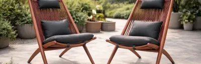 Zwei rote Stühle mit schwarzen Kissen auf einer Terrasse. Im Hintergrund Pflanzen. Die Stühle sind aus Holz mit gespannten Seilen.