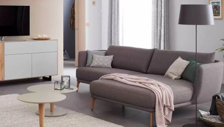 Wohnzimmer mit grauem Sofa, Kissen und Decke. Zwei runde Tische, TV-Kommode und Stehlampe sind auch zu sehen. Durchgang zu anderem Raum im Hintergrund.