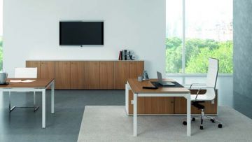 Ein Büro mit einem großen Fenster. Zwei Schreibtische stehen im Raum, einer davon mit Laptop und Bürostuhl. An der Wand hängt ein Flachbildfernseher über einer Kommode.