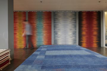 Ein Innenraum mit Teppichen, darunter ein blauer Teppich mit geometrischen Mustern, der im Vordergrund liegt. Im Hintergrund sind abstrakte Wandteppiche in Rot-, Grau- und Orangetönen zu sehen. Eine verschwommene Figur geht vorbei.