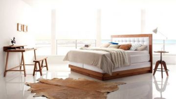 Ein helles Schlafzimmer mit Holzbett, Schreibtisch und Meerblick. Auf dem glänzenden Boden liegt ein Kuhfellteppich. Eine Lampe steht neben dem Bett. Der Raum ist modern und minimalistisch.