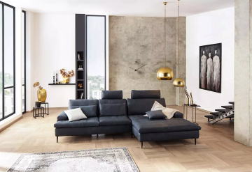 Ein modernes Wohnzimmer mit grauer Eckcouch, Holzboden, goldenen Hängelampen und abstrakten Kunstwerken. Große Fenster lassen viel Licht in den Raum. Ein Teppich liegt vor der Couch.